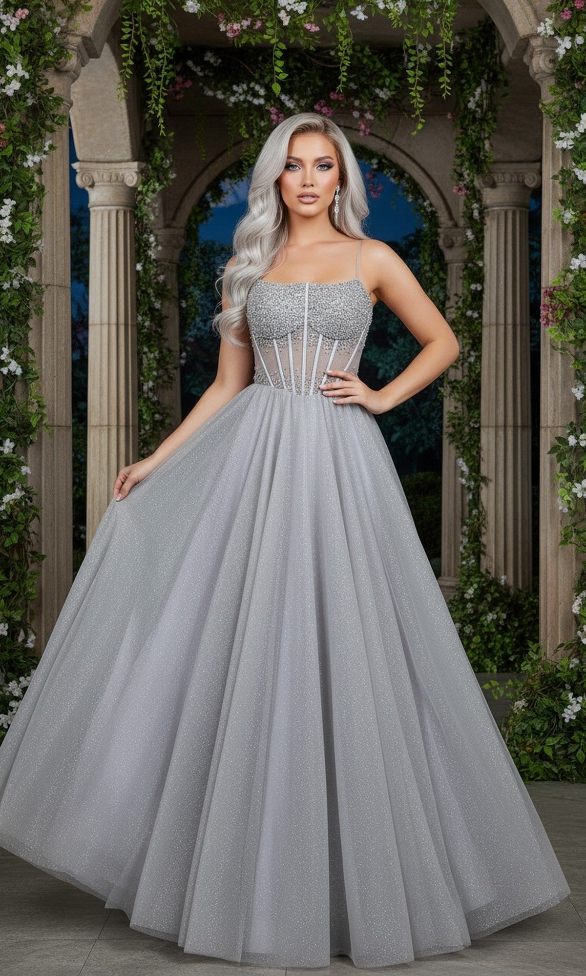 Wearing the Cinderella Couture 8459J long prom dress in silver, a woman stands before stone columns and greenery, holding out her sparkling gown's skirt.